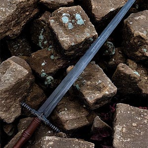 May include: A long, dark grey sword with a brown handle and ornate crossguard rests on a pile of rough, brown stones. The blade has etched markings. The handle is wrapped in brown leather. The crossguard has a decorative design.