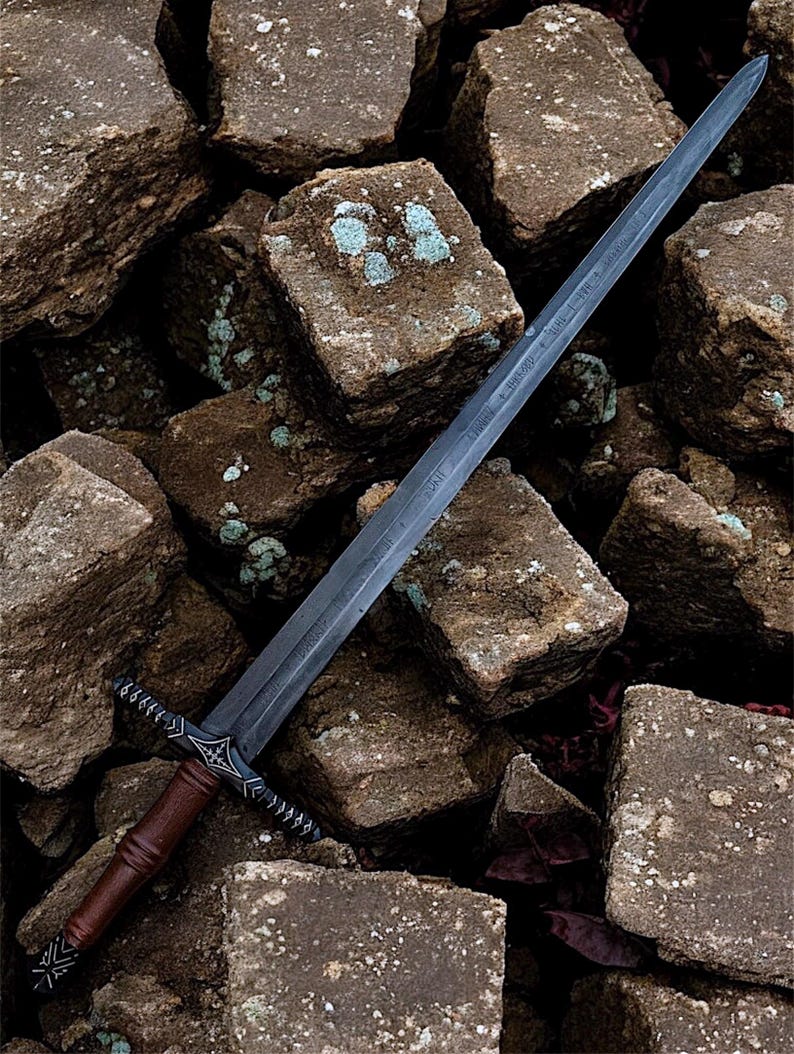 May include: A long, dark grey sword with a brown handle and ornate crossguard rests on a pile of rough, brown stones. The blade has etched markings. The handle is wrapped in brown leather. The crossguard has a decorative design.