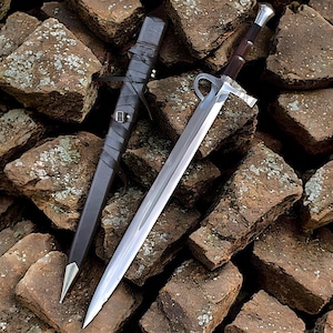 May include: A long, silver sword with a brown handle and a black sheath. The sword features a curved crossguard and a pointed tip. The sheath is made of leather and has a metal tip. The sword and sheath are on a pile of brown rocks.