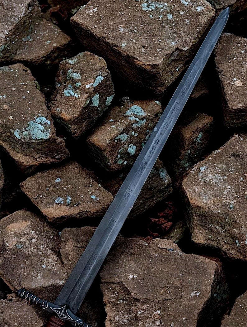 May include: A long, dark grey sword with an ornate hilt and a blade inscribed with text. The sword is displayed against a backdrop of rough, brown rocks with patches of blue-green lichen.