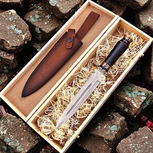 May include: A decorative dagger with a patterned blade and a dark handle, presented in a wooden box with a leather sheath. The sheath is brown and the box is lined with straw.