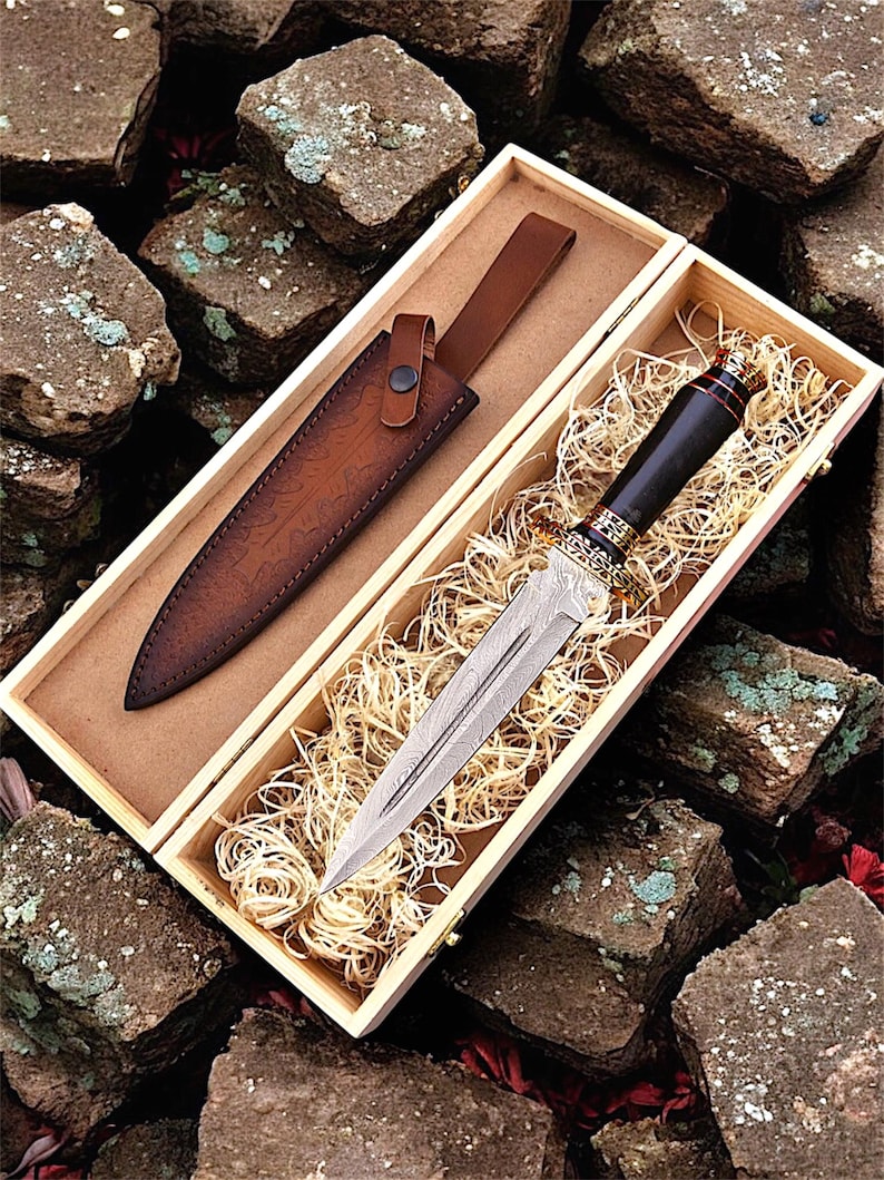 May include: A decorative dagger with a patterned blade and a dark handle, presented in a wooden box with a leather sheath. The sheath is brown and the box is lined with straw.