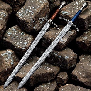 Puede incluir: Dos espadas decorativas con guardas plateadas ornamentadas y hojas estampadas descansan sobre una pila de rocas. Una espada tiene un mango envuelto en cuero marrón, y la otra tiene un mango envuelto en cuero azul. Las hojas tienen una apariencia texturizada detallada.