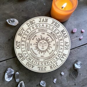 May include: A wooden divination board with engraved text and a sun design. The board features months, days of the week, numbers, and the alphabet. A lit orange candle and crystals are also present, suggesting a spiritual or mystical theme.