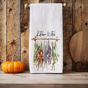 Puede incluir: Toalla de cocina blanca con tejido de gofres con el texto "Kitchen Witch" y una ilustración de acuarela de hierbas colgantes. Una pequeña calabaza naranja está sobre una superficie de madera frente a un fondo de madera.