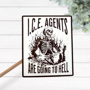 May include: A rectangular black and white sign with the text "I.C.E. AGENTS ARE GOING TO HELL". The sign features a skeleton engulfed in flames. The sign is propped up by a wooden stick.