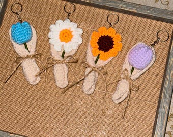 Op de afbeelding: Vier handgemaakte gehaakte bloemenboeket sleutelhangers. Elke sleutelhanger heeft een andere bloem: een blauwe tulp, een witte madelief, een oranje zonnebloem en een paarse tulp. De boeketten zijn verpakt in beige garen en vastgebonden met touw.