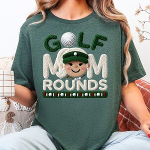 May include: A dark green t-shirt with the words "GOLF MOM ROUNDS" in a playful, embroidered style. The design includes a golf ball and a smiling cartoon face wearing a golf visor. The shirt is a casual, comfortable style.