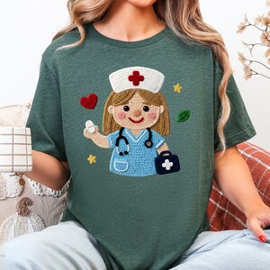 以下が含まれることがあります： 緑色のTシャツ。かぎ針編みの看護師のデザイン。看護師は青いユニフォーム、赤い十字の白い帽子を着用し、薬を持っています。聴診器、医療バッグ、装飾的な要素がデザインを完成させています。
