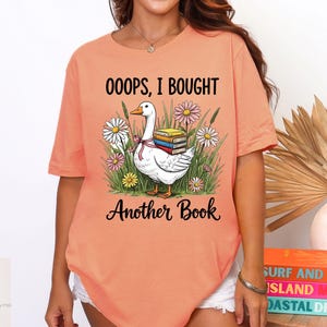 Könnte beinhalten: Pfirsichfarbenes T-Shirt mit dem Text "OOOPS, I BOUGHT Another Book". Die Grafik zeigt eine weiße Gans, die Bücher trägt, umgeben von Blumen. Das Shirt ist ein lässiger Stil.