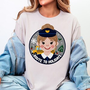 Könnte beinhalten: Beiges T-Shirt mit einem kreisförmigen Aufdruck, der eine Cartoon-Busfahrerin mit einem Bus, einer Schneeflocke und einem Weihnachtsbaum zeigt. Der Text "ROUTE TO HOLIDAYS" ist um die Grafik herum aufgedruckt. Das Shirt wird von einer Person getragen.