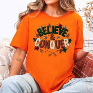 以下が含まれることがあります： 秋をテーマにしたデザインで「BELIEVE & CONQUER」の文字がプリントされたオレンジ色の半袖Tシャツ。デザインには葉、矢、ヒョウ柄が含まれています。Tシャツはソファに座っている人が着ています。