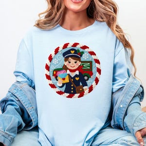May include: Light blue long-sleeve shirt featuring a circular graphic of a postal worker with a red mail truck. The graphic is surrounded by a red and white candy cane-like border. The postal worker is holding letters and wearing a navy blue uniform.