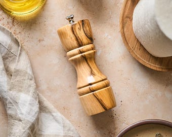 May include: Overhead shot of a rustic kitchen scene with a wooden pepper grinder, a small wooden bowl of salt with a spoon, a bottle of olive oil, a plate with cheese, a checkered cloth, and a plate with silverware. The items are arranged on a beige surface.