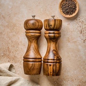 May include: Two tall, hourglass-shaped wooden pepper mills with a polished finish and visible natural grain patterns. Each mill features a small metallic knob at the top for adjustment. A small wooden bowl filled with black peppercorns is nearby.
