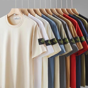 May include: A collection of short-sleeved t-shirts in various colors, including cream, white, blue, and black, hanging on wooden hangers. Each shirt features a round neck and a patch on the left sleeve.