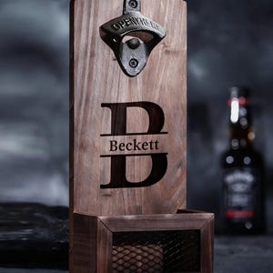 May include: A wooden bottle opener with the name "Beckett" engraved below a large letter "D". The opener is made of dark wood and has a metal bottle opener attached. A small wire mesh basket is at the bottom.