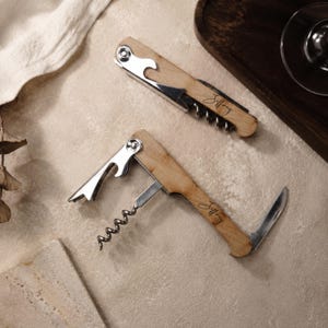 Op de afbeelding: Twee kelnerskurkentrekkers met lichthouten handvatten, elk gegraveerd met de naam Jeffrey. Eén kurkentrekker is dichtgeklapt, terwijl de andere open is en een metalen spiraal, een foliesnijder en een hefboommechanisme voor flessen toont.