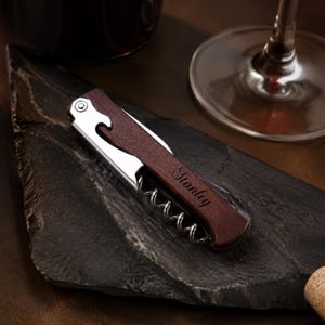 May include: A folding wine corkscrew featuring a dark brown wooden handle with the name Stanley engraved in a script font. The tool includes a polished silver metal lever, a bottle opener notch, and a spiral worm mechanism for opening wine bottles.