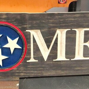 May include: A wooden sign shaped like the state of Tennessee with the word "HOME" carved into it. The sign is painted in a dark brown colour and has a blue circle with three white stars in the centre. The circle is outlined in red.