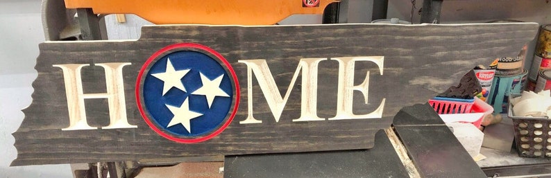May include: A wooden sign shaped like the state of Tennessee with the word "HOME" carved into it. The sign is painted in a dark brown colour and has a blue circle with three white stars in the centre. The circle is outlined in red.