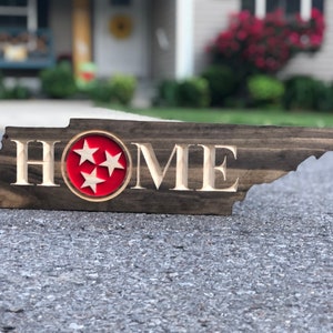 May include: A wooden sign shaped like the state of Tennessee with the word "HOME" carved into it. The sign has a red circle with three white stars in the centre, representing the state flag of Tennessee.
