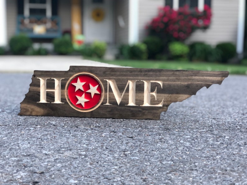 May include: A wooden sign shaped like the state of Tennessee with the word "HOME" carved into it. The sign has a red circle with three white stars in the centre, representing the state flag of Tennessee.