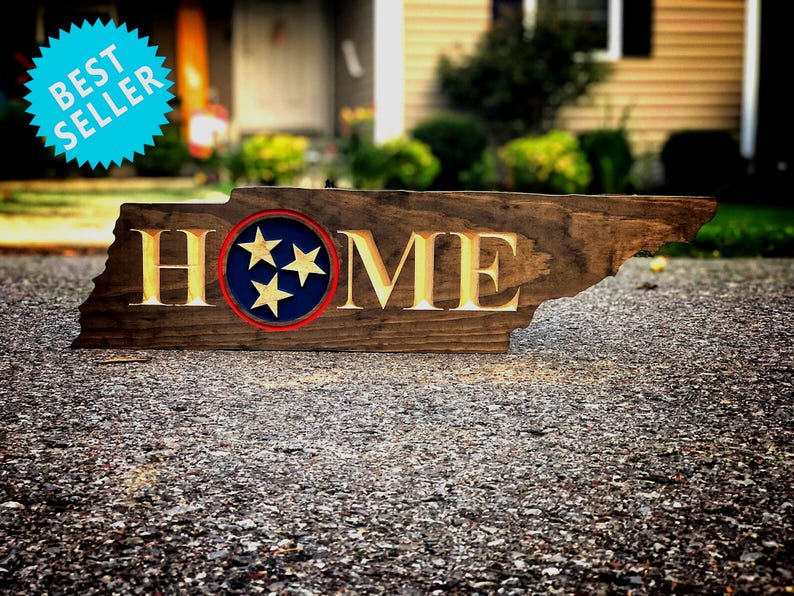 May include: A wooden sign shaped like the state of Tennessee with the word "HOME" carved into it. The sign is painted brown with a blue circle in the centre featuring three white stars.