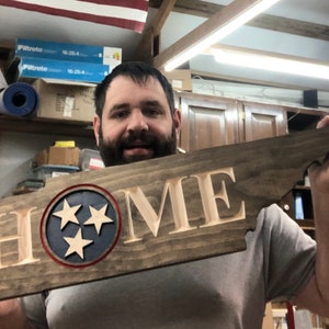 May include: A wooden sign shaped like the state of Tennessee with the word "HOME" carved into it. The sign features a blue circle with three white stars, representing the state flag of Tennessee.