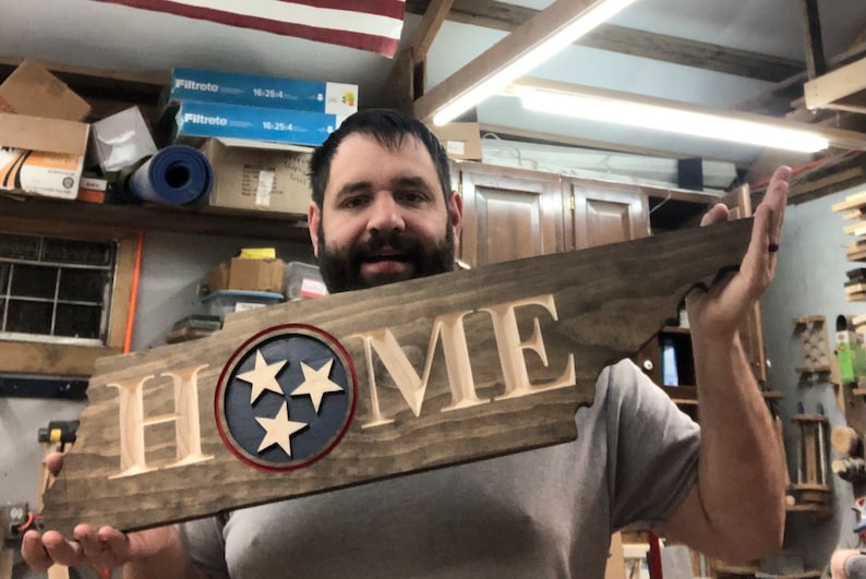 May include: A wooden sign shaped like the state of Tennessee with the word "HOME" carved into it. The sign features a blue circle with three white stars, representing the state flag of Tennessee.
