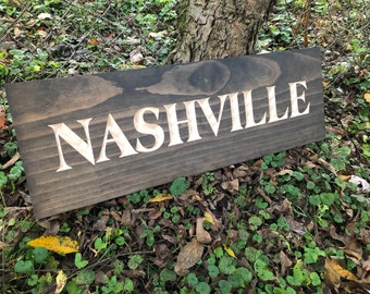 May include: A rectangular wooden sign with the word "NASHVILLE" carved into it. The letters are a light color, contrasting with the dark stained wood. The sign is displayed outdoors.