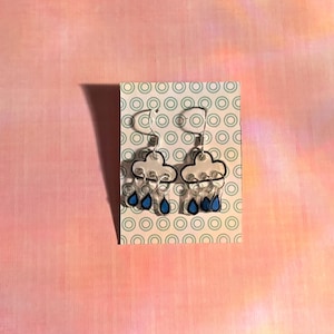 Peut inclure: Boucles d'oreilles en forme de nuage avec des gouttes de pluie bleues. Les boucles d'oreilles ont un contour blanc et sont présentées sur une carte avec un motif circulaire vert et blanc. Le fond est un dégradé rose et orange.