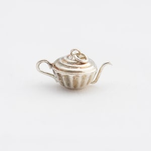 May include: A small silver-colored metal charm shaped like a miniature teapot. The charm features a rounded body with vertical fluted detailing, a curved handle, a thin spout, and a small circular jump ring attached to the top lid for hanging.