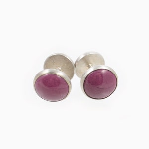 May include: A pair of silver-toned cufflinks with round, polished, magenta-colored stones. The cufflinks are set against a white background, highlighting their design and color.