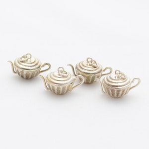 May include: Four small, silver-colored teapot charms with detailed designs. Each charm has a handle, spout, and a lid with a loop. The teapots are arranged on a white background.