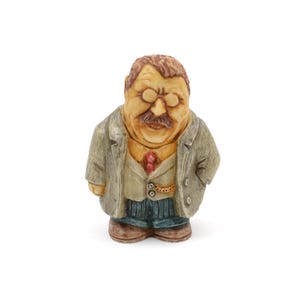 May include: A carved wooden figurine of a man wearing a suit and glasses. The figure has a mustache and is wearing a gray coat, a light-colored vest, a red tie, and blue pants. The figurine is standing with its hands in its pockets.