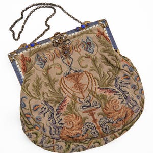 May include: An antique tapestry handbag with a metal chain strap. The bag features a beige background with a detailed floral and heraldic design in shades of brown, green, and blue. The frame is accented with blue beads.