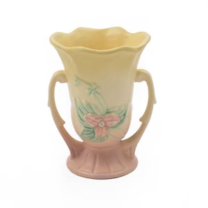 May include: A ceramic vase featuring a scalloped rim and two curved handles. The body displays a gradient color scheme transitioning from pale yellow at the top to a soft pink base, accented with a raised relief of a pink flower and green leaves.