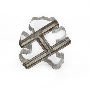 May include: A silver-colored metal cookie cutter featuring a multi-lobed, flower-like shape with a central crossbar handle. The cutter is constructed from thin, bent metal strips with visible seams and a weathered, industrial metallic finish.
