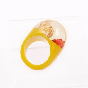 May include: A unique, yellow resin ring with a clear, oval-shaped top. The top encases a small seashell and red floral elements. The ring's design is modern and artistic, suitable for adding a touch of nature-inspired flair to any outfit.