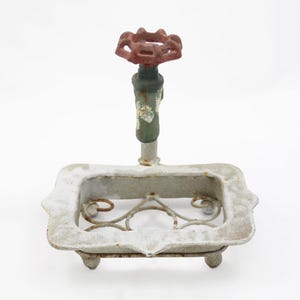 May include: A white metal soap dish featuring a rectangular frame with a decorative scrollwork base and four small feet. A vertical pipe attachment extends from the center, topped with a red, multi-sided manual water valve handle.