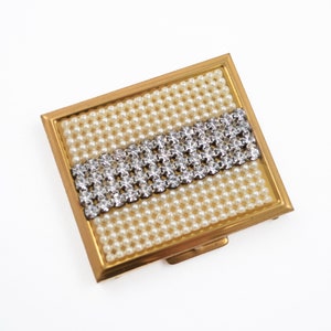 May include: A vintage square compact with a gold-tone frame. The center features a band of sparkling rhinestones set against a background of small, cream-colored faux pearls. The compact is open, revealing the interior.