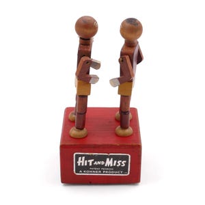 May include: A vintage wooden toy featuring two articulated figures standing on a red base. The figures are brown with tan and silver accents. The base has a label that reads "Hit and Miss".