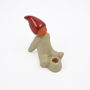 May include: A ceramic candle holder shaped like a seated figure wearing a glossy red pointed hat. The figure has a matte beige body and an orange face, with a circular opening for a candle integrated into the base near the figure's legs.