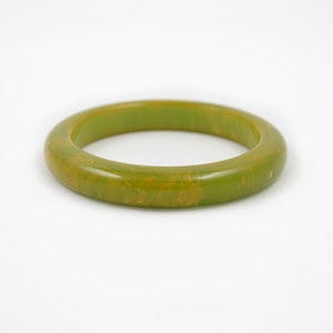 May include: A round, olive green bangle bracelet. The bracelet has a smooth, polished surface with subtle variations in color, including hints of yellow and brown. This vintage-style bracelet is a classic accessory.