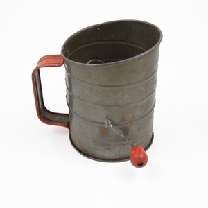 May include: Vintage metal flour sifter with a red handle and knob. The cylindrical sifter has a gray finish with a red handle and knob. The handle is a curved, rectangular shape. The sifter is designed for sifting flour and other dry ingredients.