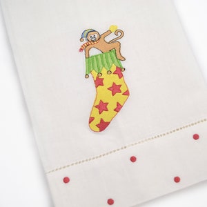 May include: A white hand towel featuring an embroidered design of a brown monkey wearing a blue and yellow striped hat and a red and white striped scarf, emerging from a yellow Christmas stocking with red stars and a green scalloped cuff.