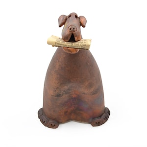 May include: A brown ceramic dog figurine with a smooth, rounded body and floppy ears. The dog is holding a small, rolled-up piece of newspaper in its mouth. The sculpture features a dark, mottled finish and is shaped with a wide base and two front paws.