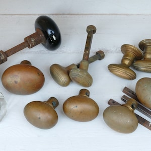 May include: A collection of antique door knobs in various styles and finishes. The knobs include black glass, clear glass, and brass, with some showing signs of age and wear. The hardware is arranged on a white surface.