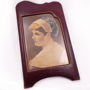 May include: An antique portrait of a woman in profile, framed in a burgundy leather-bound frame with gold trim. The portrait features a woman with blonde hair, wearing a white shawl and a blue and lace head covering.